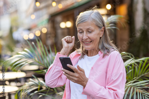 Senior woman outside, fist pumped in victory, smiling at her smartphone, capturing the joy of good news.