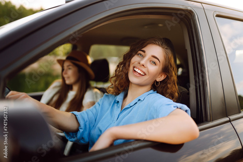 Two trendy attractive young woman singing along to the music as they drive along in the car through town. Beautiful female friends in the car enjoy a car trip together. © maxbelchenko