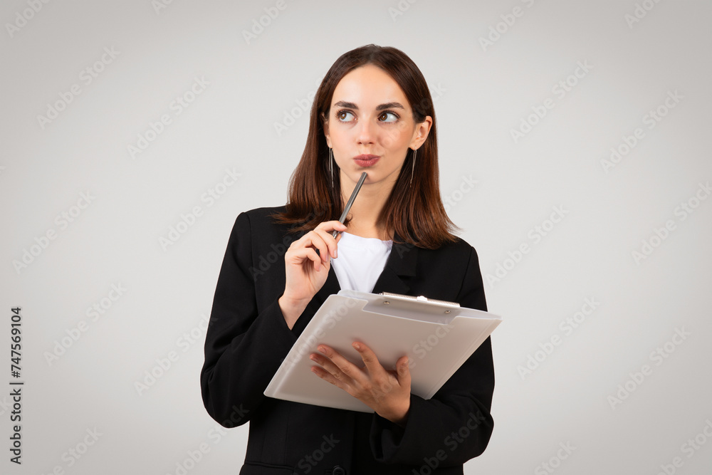 Thoughtful businesswoman holding a clipboard pensively touches her chin with a pen Thoughtful businesswoman holding a clipboard pensively touches her chin with a pen