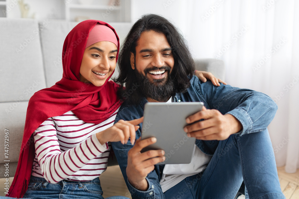 Smiling muslim couple with digital tablet relaxing on floor at home Smiling muslim couple with digital tablet relaxing on floor at home