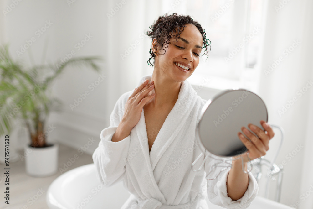 Pretty young black woman in bathrobe looking at mirror and smiling, touching neck and enjoying her moisturised skin, sitting in bathroom, copy space Pretty young black woman in bathrobe looking at mirror and smiling, touching neck and enjoying her moisturised skin, sitting in bathroom, copy space