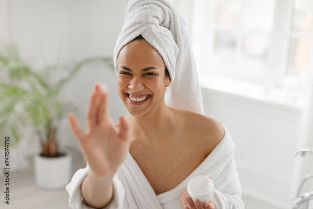 Playful black woman using cream after shower and having fun, stretching hand to camera, sitting in bathroom at home. Facial skincare cosmetics concept