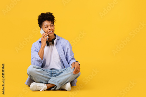 Happy black male student talking on phone with headphones on yellow background © Prostock-studio