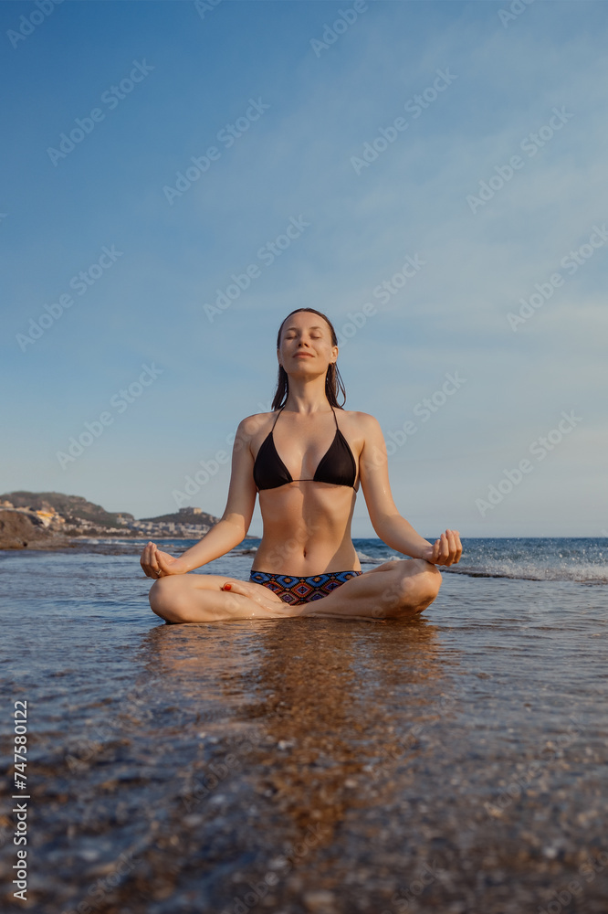 A woman in a swimsuit finds tranquility in meditation on the sunlit beach, embracing the serene sea ambiance