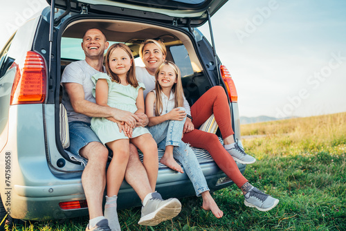 Portrait of Happy young couple with little daughters sitting inside car trunk during auto trop. They are smiling, laughing and chatting. Family values, traveling concept. © Soloviova Liudmyla