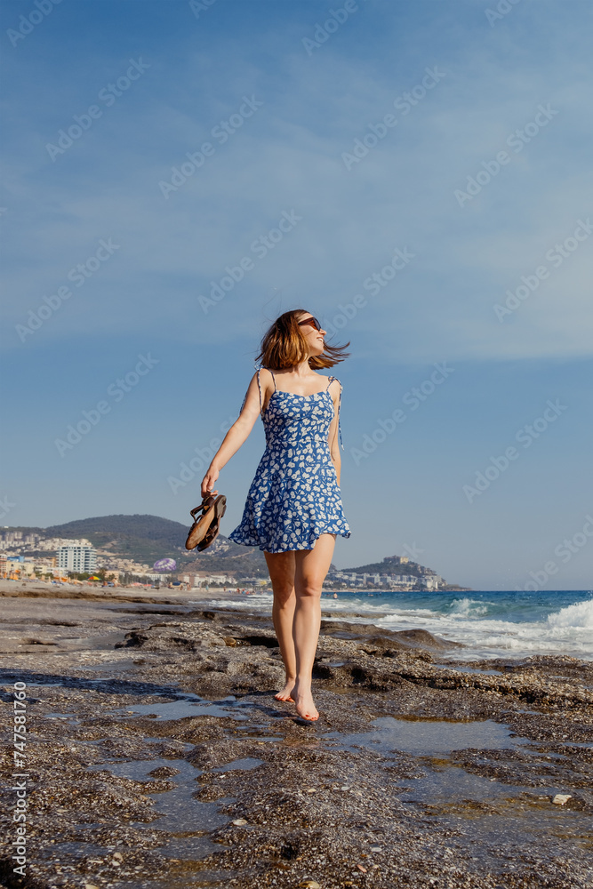 A woman enjoys a leisurely walk along the rocky beach, feeling the summer breeze in her flowing dress