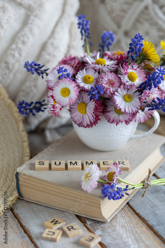 Colourful bouquet with blue flowers  meadow daisy in a cup on the table. Old book  straw hat  countryside home decor. Postcard for the holidays. Word summer written in wooden letters. Calendar