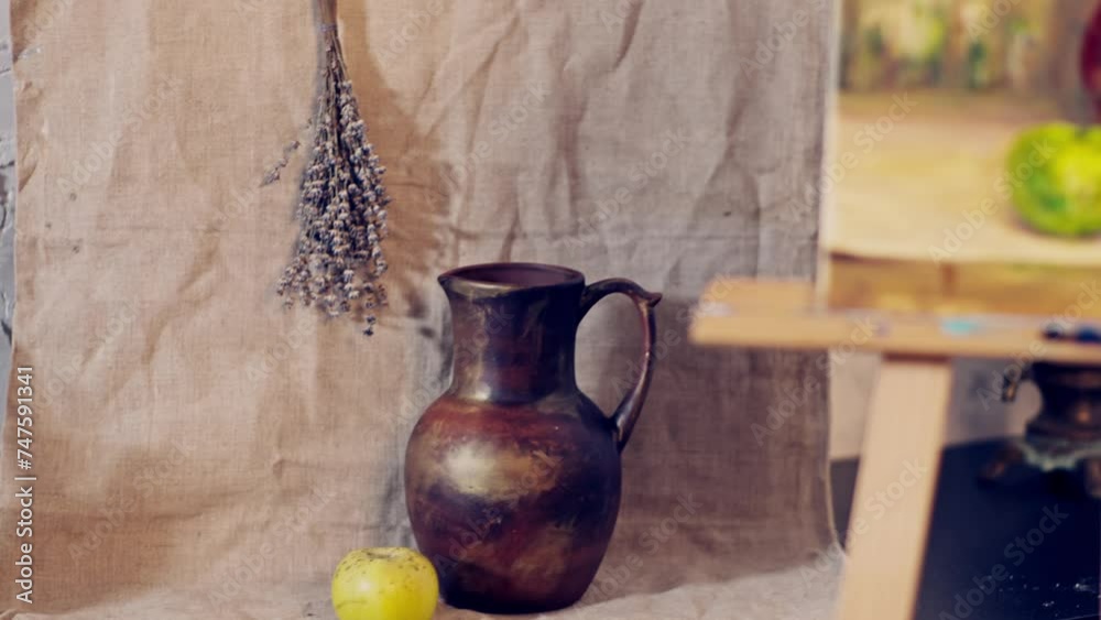 Still life objects and readymade painting in the artist’s studio. Jug, apple and dry flowers on the table. Painting in warm colors on the easel. Still life objects and readymade painting in the artist’s studio. Jug, apple and dry flowers on the table. Painting in warm colors on the easel.
