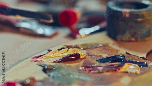 Artist mixing paints by a palette knife on the wooden palette. Red and yellow oil paints on the artist’s palette. Artist studio. 