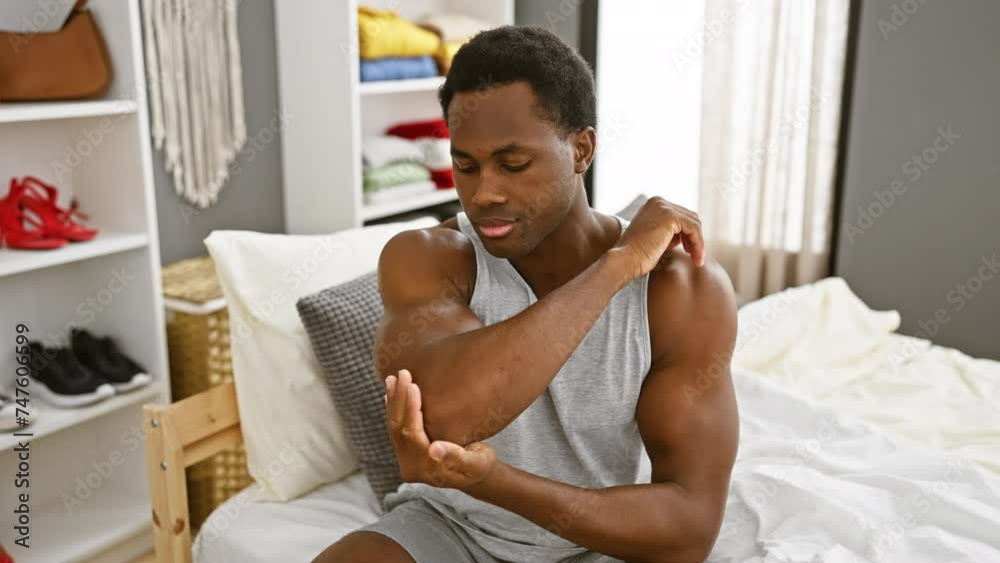 A young adult black man applying cream on his elbow while sitting on a bed indoors with a blurred shelf in the background. A young adult black man applying cream on his elbow while sitting on a bed indoors with a blurred shelf in the background.