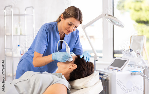 Professional positive female aesthetician administering high frequency ultrasound facial procedure on mature woman in cosmetology clinic. Modern technologies for skin rejuvenation