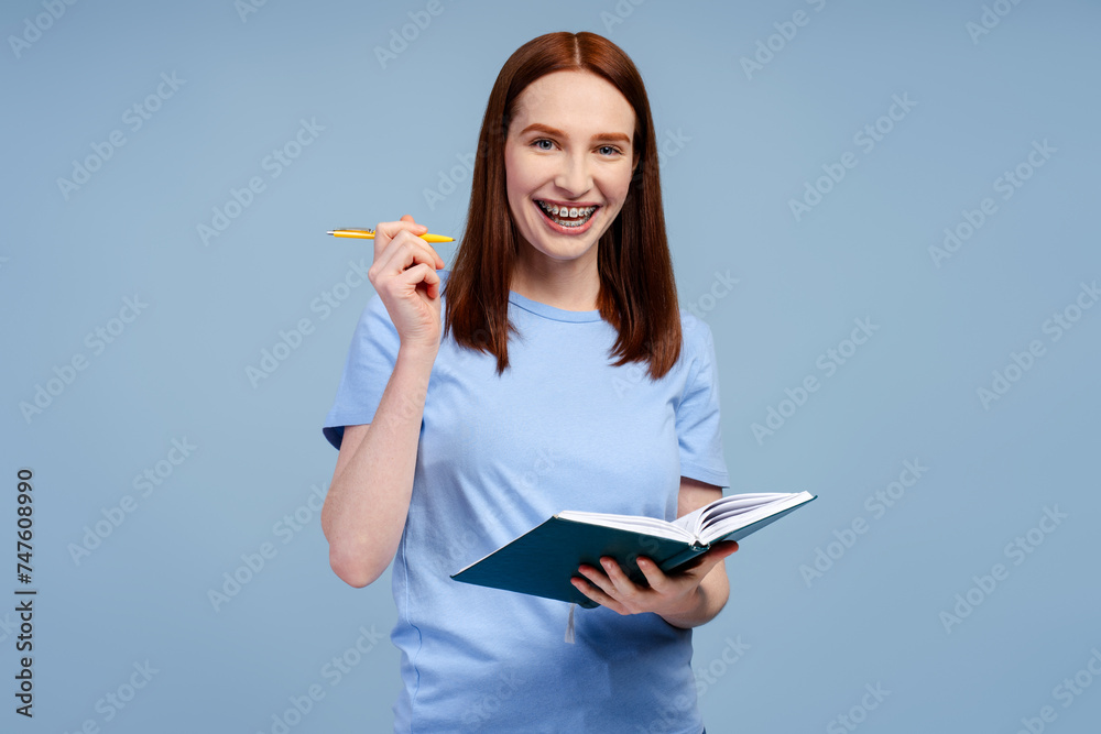 Smiling university ginger student wearing blue t shirt holding open book isolated on blue background Smiling university ginger student wearing blue t shirt holding open book isolated on blue background