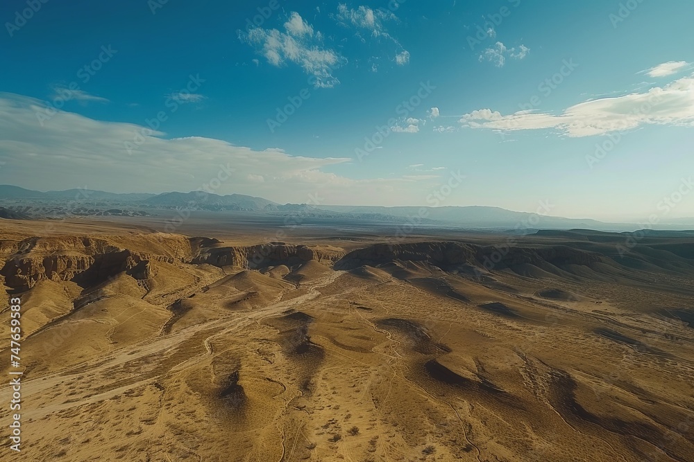 Aerial view shows a vast desert landscape with mountains towering in the background. The arid terrain stretches out under the clear blue sky. Generative AI