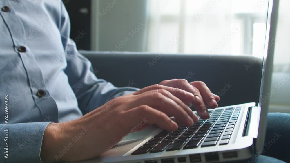 Man works on a laptop while sitting on the couch at home.Close-up young man working on his laptop at home, bokeh view of a business man's hands busy using a laptop, typing on a computer, sitting sofa