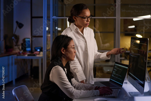 Ethnically diverse female programmers fixing code at night in modern office