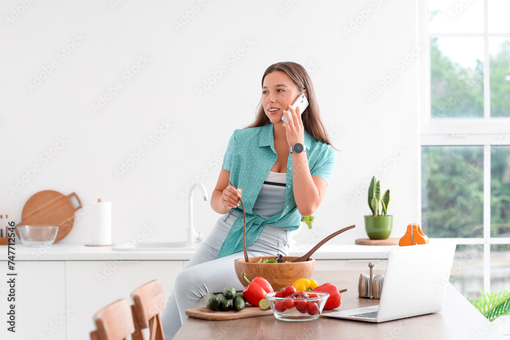 Young woman talking by mobile phone while preparing vegetable salad in kitchen