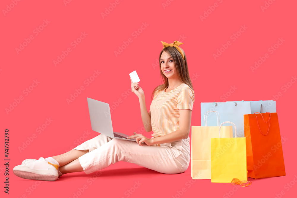 Beautiful happy stylish young woman with shopping bags, credit card and laptop sitting on red background Beautiful happy stylish young woman with shopping bags, credit card and laptop sitting on red background