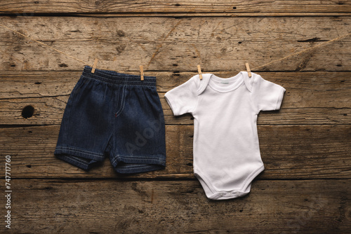 A pair of baby denim shorts and a white onesie are pinned to a clothesline against a rustic wooden b