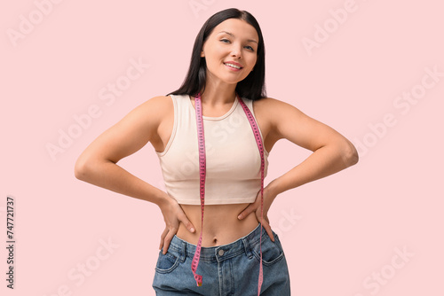Beautiful young happy woman in loose jeans with measuring tape on pink background. Weight loss concept © Pixel-Shot