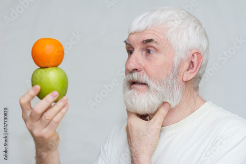 A gray-haired elderly man holds a tangerine and a green apple in his hands