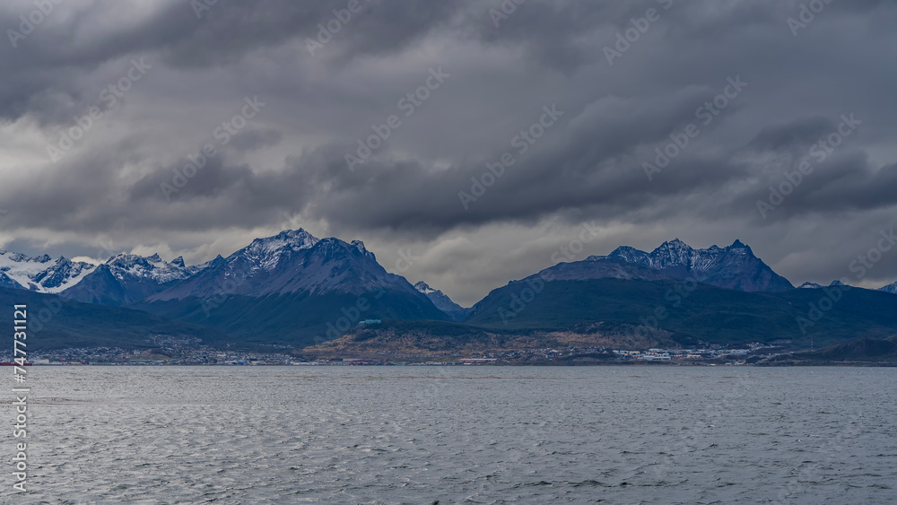 A beautiful snow-capped mountain range of the Andes against a cloudy sky. View from the Beagle Canal. The town houses of Ushuaia are visible on the coast. Tierra del Fuego Archipelago. Argentina