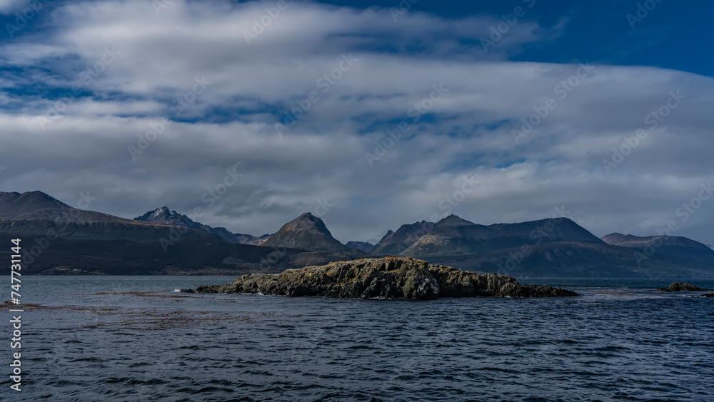 Small rocky islets in the Beagle Channel. Ripples on the blue water. The coastal mountain range of the Andes against the background of sky and clouds. Tierra del Fuego Archipelago. Argentina