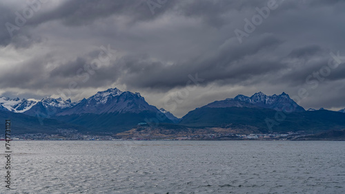 A beautiful snow-capped mountain range of the Andes against a cloudy sky. View from the Beagle Canal. The town houses of Ushuaia are visible on the coast. Tierra del Fuego Archipelago. Argentina