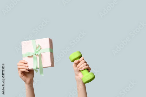 Female hands with dumbbell and gift box for International Women's Day on grey background