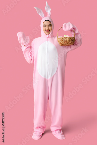 Beautiful young woman in bunny costume holding wicker basket with Easter cake and eggs on pink background