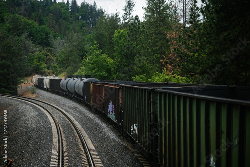A long train with a mixed manifest of boxcars and tankcars, showing freight of goods such as fossil fuels and phosphate. The train is in a forest, alluding to the vast distances of American transport. photo