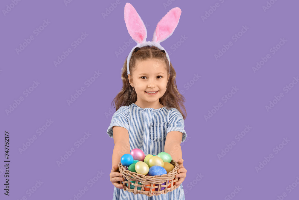 Cute little girl in bunny ears holding wicker basket with Easter eggs on purple background