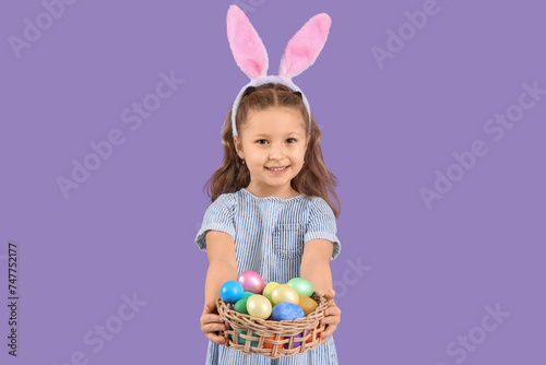 Cute little girl in bunny ears holding wicker basket with Easter eggs on purple background