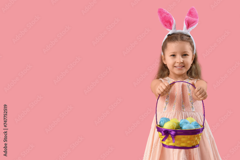 Cute little girl in bunny ears holding wicker basket with Easter eggs on pink background
