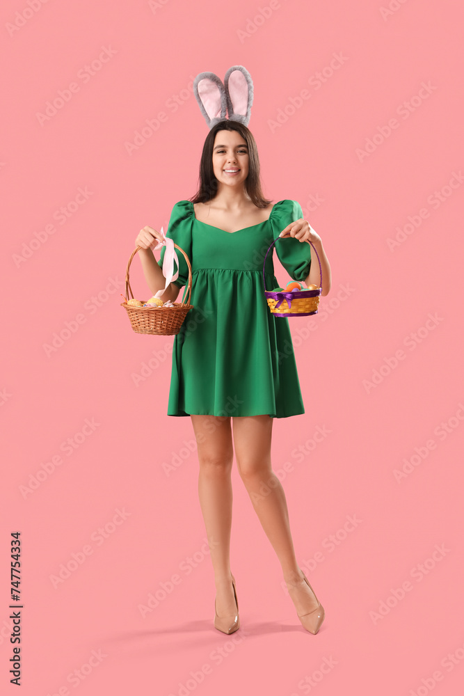 Beautiful young woman in bunny ears holding wicker baskets with Easter eggs on pink background