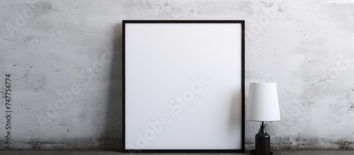 A blank black and white picture frame hangs on a cement wall in an interior room. Next to the frame, a white lamp provides illumination.