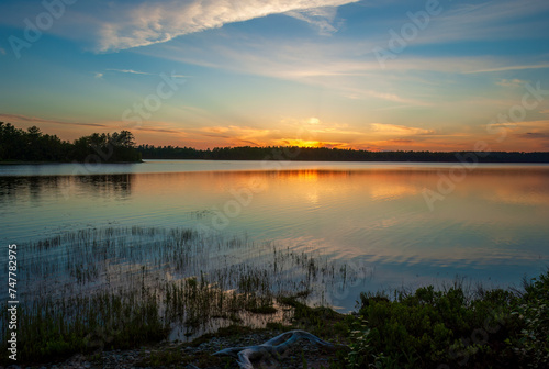 Serene sunset over Graham Lake, in Maine, USA. Colorful sky reflected on rippled waters. Aquatic vegetation on the shoreline. © Rasvan