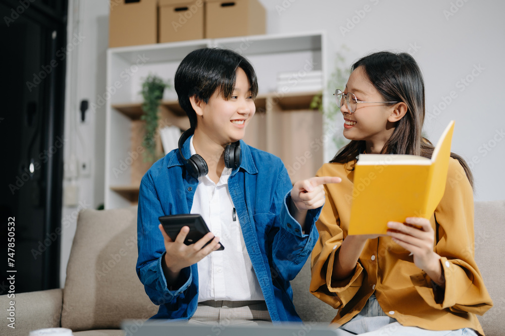 Obraz premium Two Attractive young Asian female college students working on the school project using laptop computer and tablet together, enjoy talking and snacking on sofa