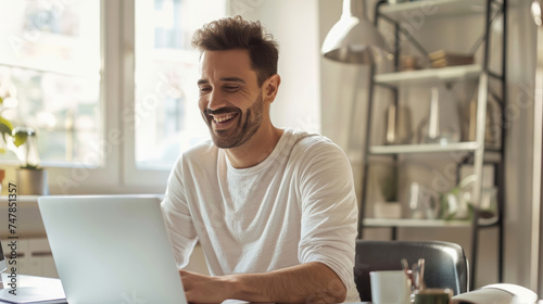 Smiling man in a white t-shirt using a laptop at a bright home office setup