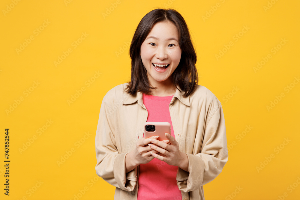 Young surprised shocked woman of Asian ethnicity wear pink t-shirt beige shirt pastel casual clothes hold in hand use mobile cell phone isolated on plain yellow background studio. Lifestyle portrait. Young surprised shocked woman of Asian ethnicity wear pink t-shirt beige shirt pastel casual clothes hold in hand use mobile cell phone isolated on plain yellow background studio. Lifestyle portrait.