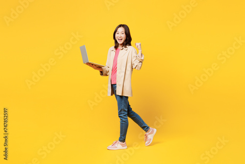 Full body young IT woman of Asian ethnicity she wear pink t-shirt beige shirt pastel casual clothes hold use work on laptop pc computer isolated on plain yellow background studio. Lifestyle portrait. © ViDi Studio