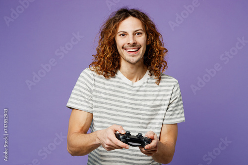Young smiling fun man he wears grey striped t-shirt casual clothes hold in hand play pc game with joystick console isolated on plain pastel light purple background studio portrait. Lifestyle concept.