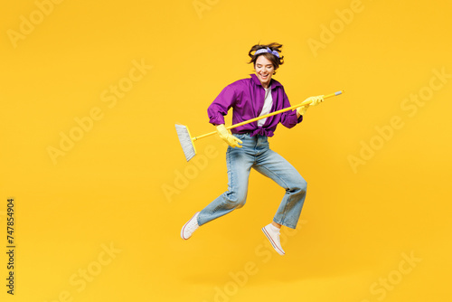 Full body young fun woman wear purple shirt casual clothes do housework tidy up jump high hold brush broom pov play guitar jump high isolated on plain yellow background studio. Housekeeping concept.