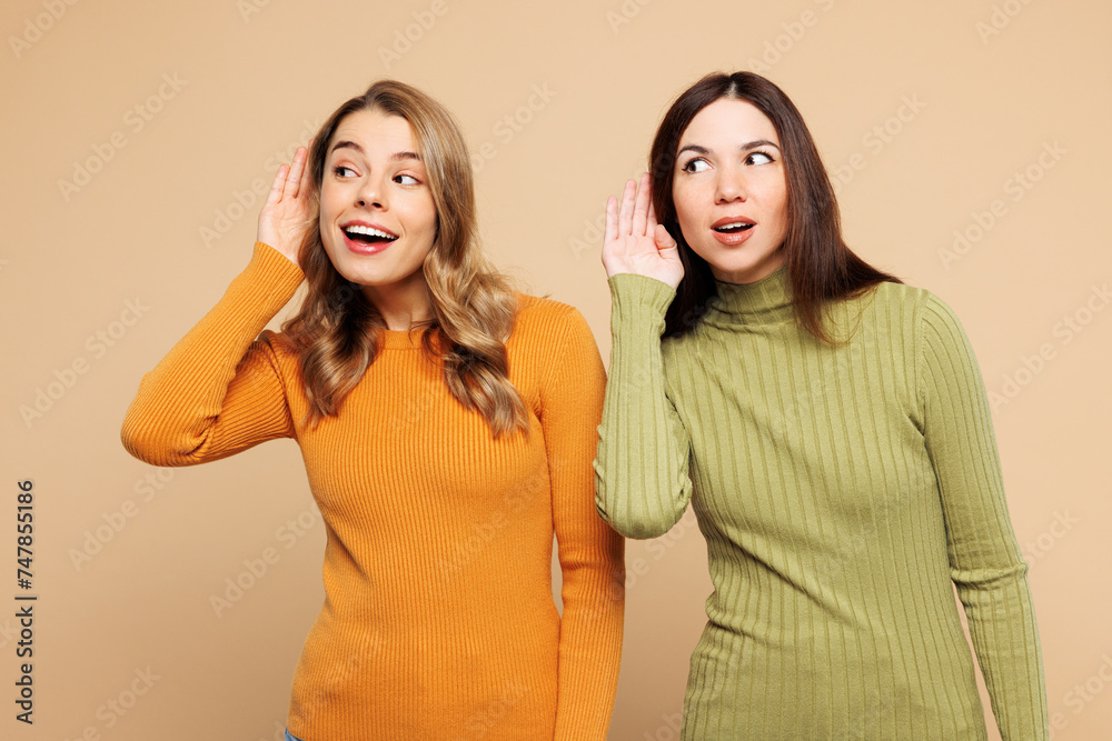 Young curious nosy friend two women they wear orange green shirt casual clothes together try to hear you overhear listening intently isolated on plain pastel light beige background. Lifestyle concept Young curious nosy friend two women they wear orange green shirt casual clothes together try to hear you overhear listening intently isolated on plain pastel light beige background. Lifestyle concept