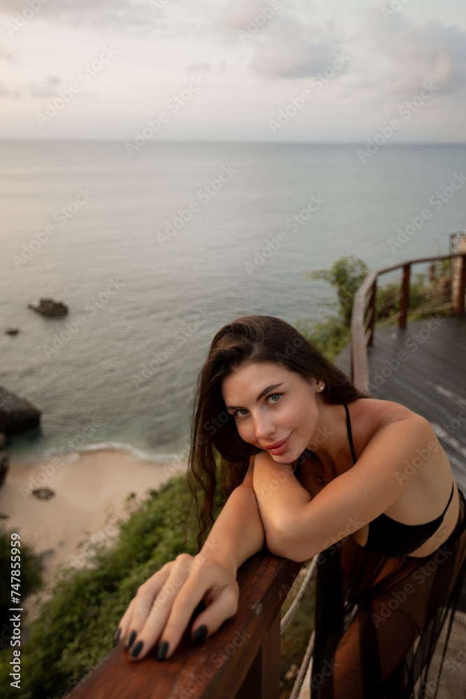 Young pretty brunette woman stand on the balcony of hotel resort with ocean sea view, look at the camera