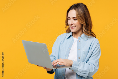 Young smart happy IT woman she wear blue shirt white t-shirt casual clothes hold use work on laptop pc computer chatting online isolated on plain yellow background studio portrait. Lifestyle concept.