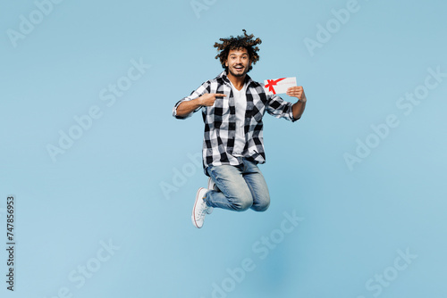 Full body young Indian man wears shirt white t-shirt casual clothes jump high hold point finger on gift certificate coupon voucher card for store isolated on plain blue background. Lifestyle concept.