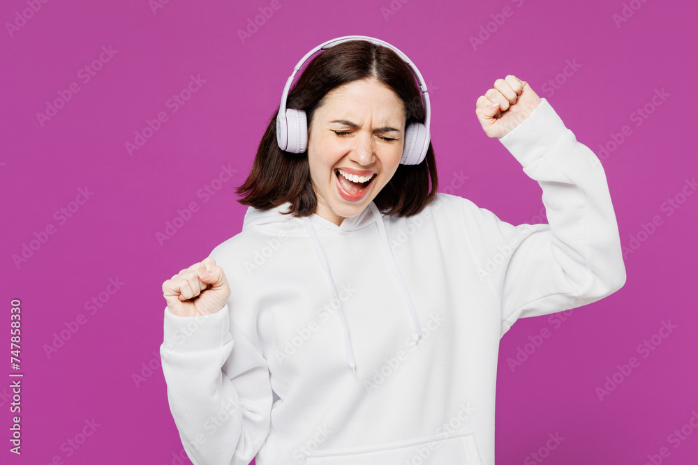Young cool smiling happy Caucasian woman she wear light hoody casual clothes listen to music in headphones raise up hands isolated on plain purple color background studio portrait. Lifestyle concept. Young cool smiling happy Caucasian woman she wear light hoody casual clothes listen to music in headphones raise up hands isolated on plain purple color background studio portrait. Lifestyle concept.