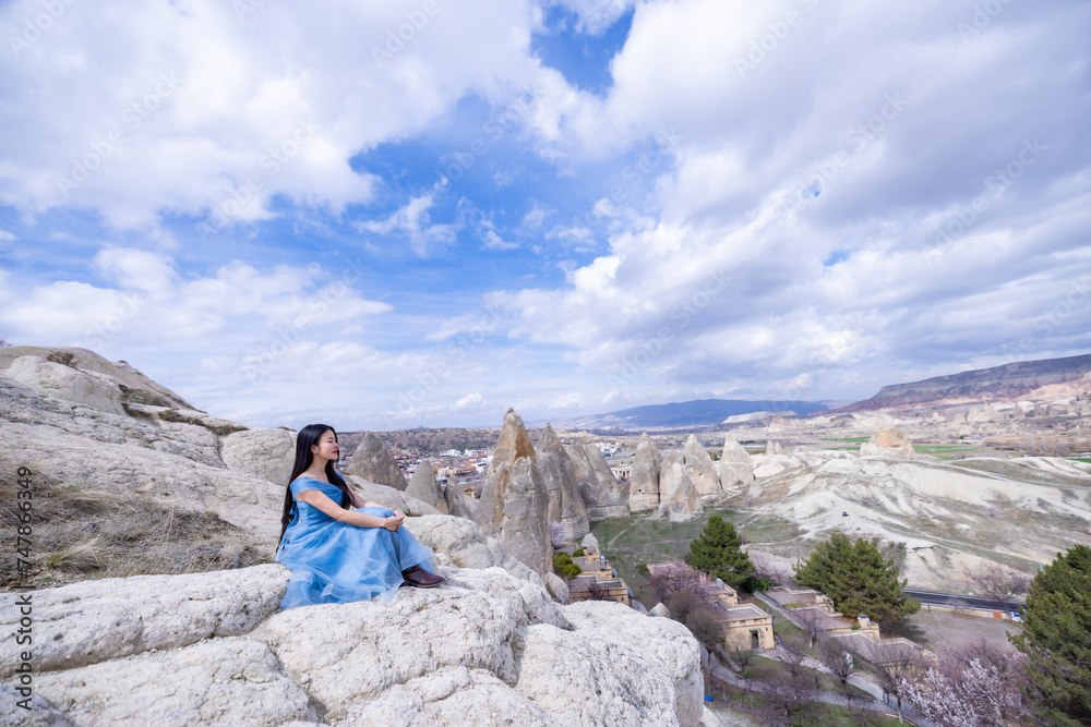 Fototapeta premium Beautiful woman in blue dress on Turkey rocks mountain.Cheerful woman relax outdoor with beautiful sky and pink flowers in holiday vacation.Woman tourist enjoy amazing mountain view.Travel and freedom