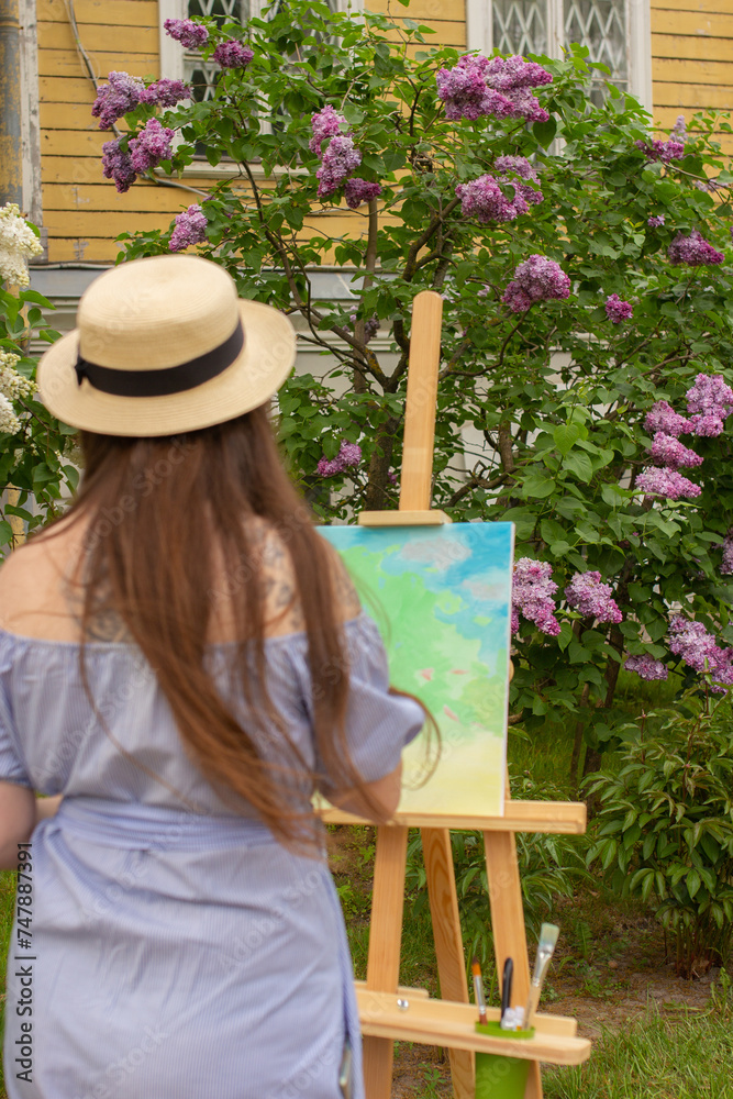 Female artist painting on plein air in spring lilac garden. Woman in hat with wooden easel and canvas standing outdoor
