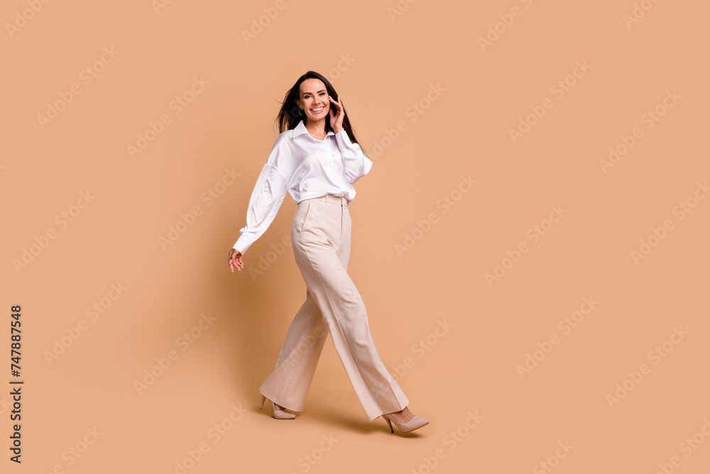 Full length photo of pretty positive woman wear white shirt wind blowing walking empty space isolated beige color background Full length photo of pretty positive woman wear white shirt wind blowing walking empty space isolated beige color background